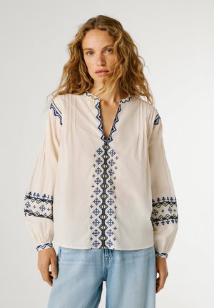 Mujer con cabello rubio rizado que lleva una blusa crema con bordados geométricos en azul y negro y jeans azul claro, de pie contra un fondo liso.