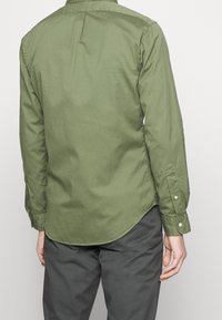 Olivgrön långärmad skjorta med knapp-ner krage, bakveck och rundad nederkant. Tyget ser slätt ut och har en lätt textur.