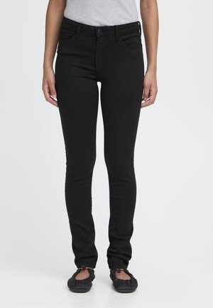 Frau trägt eng anliegende schwarze Jeans, graues ärmelloses Oberteil und schwarze Ballerinas, steht vor einem einfarbigen Hintergrund.