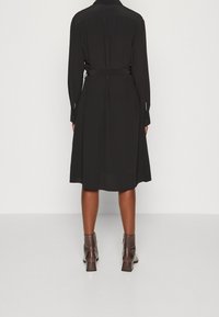Abito nero a maniche lunghe con vita cinturata, silhouette a forma di A e chiusura a bottoni ai polsini. Il tessuto appare liscio e leggero.