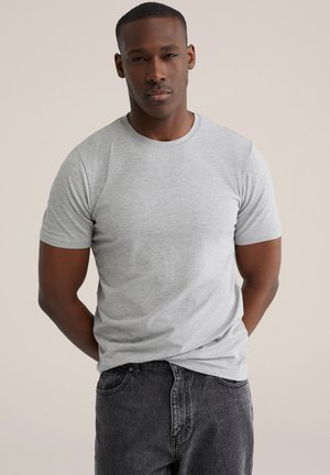 Jonge man met een donkere huidkleur, gekleed in een effen lichtgrijs T-shirt met korte mouwen en donkere spijkerbroek, staand met zijn handen achter zijn rug.