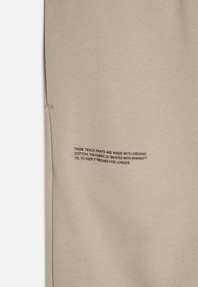 Pantalones deportivos beige hechos de algodón orgánico con una textura suave. Presentan información de cuidado impresa en negro a lo largo de la costura lateral.