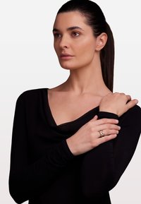 Inel de aur cu un cristal central, design stratificat. Modelul poartă un top negru cu guler tip cowl, evidențiind materialul neted și mânecile ajustate.