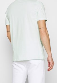 Man som bär en ljusgrön t-shirt med kort ärm och vita byxor, vänd ryggen mot kameran med taggtrådstatuer på båda överarmarna.