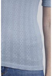 Gaiši zils īsu piedurkņu adīts tops ar teksturētu dizainu, kas satur vertikālas svītras un nelielus acs rakstus visā virsmā.