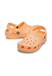 Sabots en plastique orange avec des côtés perforés, une sangle à l'arrière et des semelles texturées. Présente un logo circulaire sur le côté externe. Conception légère.