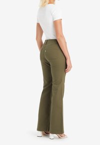 Pantalon évasé vert olive en tissu mélange de coton, avec une taille haute et deux poches arrière, accompagné d'une chemise blanche ajustée et de talons.