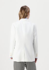 Biely blazer s prispôsobeným strihom, s jedným rozparkom a manžetami na tri gombíky. Model má na sebe čierno-biele pruhované nohavice, pohľad zozadu.