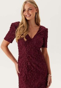 Bordeauxklänning med texturerad blommönster, korta ärmar och djup V-ringning. Slät tyg, figurnära silhuett och subtil lyster i designen.