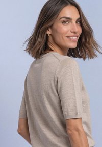 Woman with shoulder-length brown hair smiling over her shoulder, wearing a beige short-sleeve knit top and gold hoop earrings against a light gray background.
