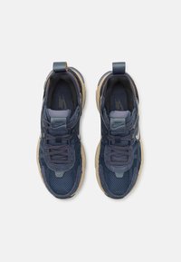 Marineblaue Sneakers mit Mesh- und Wildledertexturen, ausgestattet mit einer beigen Gummisohle und einem reflektierenden Akzent. Die Schnürsenkel verlaufen durch strukturierte Ösen.