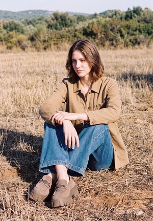 Une femme portant une veste beige, un jean bleu et des chaussures épaisses est assise dans un champ sec, avec des collines boisées derrière elle.