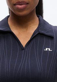 Σκούρο μπλε πουκάμισο με γιακά και διακριτικές κάθετες ρίγες, διαθέτει V-λαιμόκοψη και μικρό λευκό λογότυπο στην αριστερή πλευρά. Λείο υλικό.