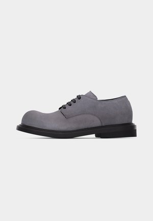 Chaussure à lacets en daim gris avec bout arrondi et semelle noire, vue de profil sur un fond clair uni.
