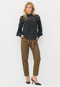 Czarna bluzka z koronkowymi detalami, długimi rękawami i falbaniastym dołem, w połączeniu z brązowymi spodniami z ekologicznej skóry o wysokim stanie i czarnymi szpilkami.