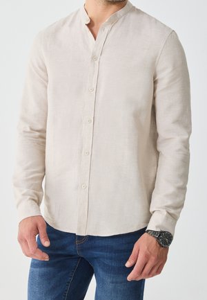 Homme portant une chemise beige à manches longues avec col mandarin et un jean bleu foncé, montre noire au poignet gauche.