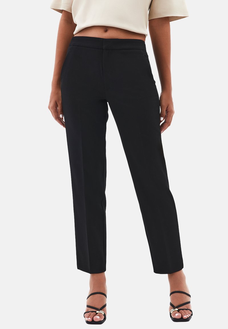 Mujer con pantalones negros ajustados hasta el tobillo y sandalias de tacón negras con tiras, de pie frente a un fondo blanco liso.