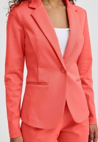 Blazer corail avec une coupe ajustée, doté d'une fermeture à un bouton, de revers crantés et de deux poches avant. Texture de tissu lisse.