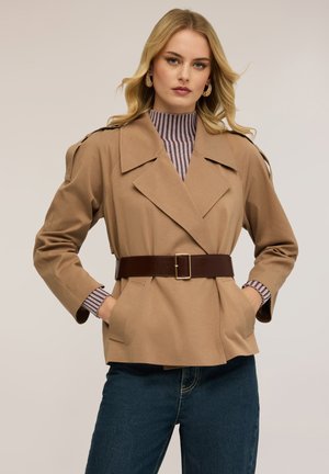 Mujer rubia con cabello ondulado que lleva una chaqueta beige con cinturón sobre un suéter de cuello alto a rayas y jeans azules, posando con las manos en los bolsillos.