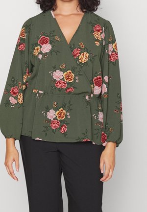 Woman wearing an olive green floral wrap blouse with long sleeves and black pants, standing against a plain light background.