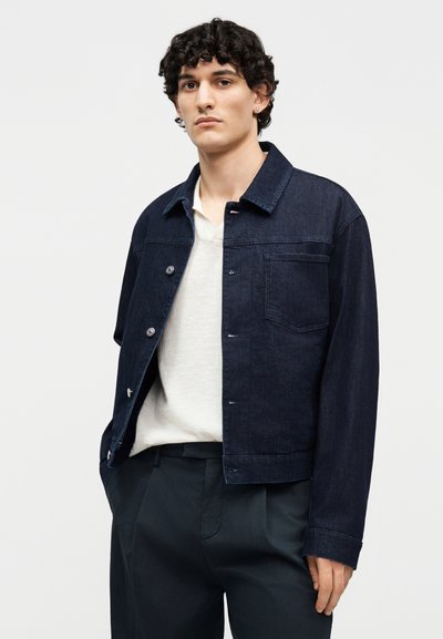 Νεαρός άνδρας με κοντά σγουρά μαλλιά, φορώντας ένα σκούρο μπλε τζιν μπουφάν πάνω από άσπρο πουκάμισο και μαύρο παντελόνι, στέκεται μπροστά σε φωτεινό φόντο.