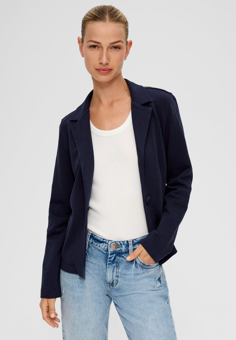 Młoda kobieta z blond włosami w granatowej marynarce, białym topie na ramiączkach i jasnoniebieskich dżinsach, stojąca z jedną ręką w kieszeni.