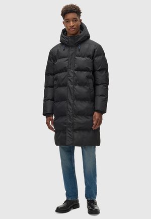 Zwarte pufferjas met een capuchon, voorzien van een ritssluiting, elastische accenten en een knielange pasvorm, gecombineerd met lichtblauwe jeans en zwarte laarzen.