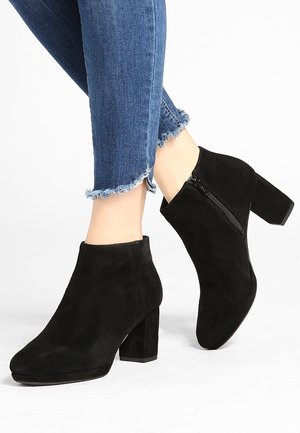 Bottines en daim noir à talon block, avec une fermeture éclair sur le côté et un bout arrondi, associées à un jean bleu à ourlets effilochés.