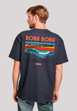Navy blue t-shirt with large retro-style "BORA BORA" graphic, featuring waves and sunset stripes in orange and teal, soft cotton texture.