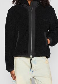 Person som bär en svart fleecejacka med dragkedja och har handen i fickan, tillsammans med ljusblå slitna jeans och ett svart snörbälte.