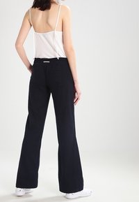 Femme portant un haut à bretelles spaghetti blanc et un pantalon noir ample avec des baskets blanches, se tenant de dos contre un fond uni.