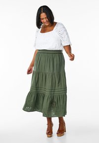 Jupe maxi verte à niveaux avec des détails brodés, associée à une blouse en dentelle blanche. Le mannequin porte des sandales à talons marron.