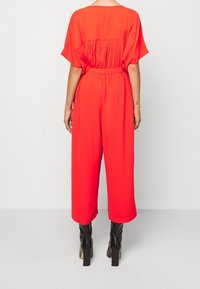 Mono rojo con un corte relajado, mangas cortas, detalle de pliegues en la parte posterior y cintura elástica. Presenta piernas ajustadas y botas de tacón negras.