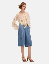 Blusa de crema translúcida con detalles de volantes, combinada con culottes de mezclilla azul que cuentan con bolsillos y un cinturón beige, complementada con sandalias de tacón marrón.