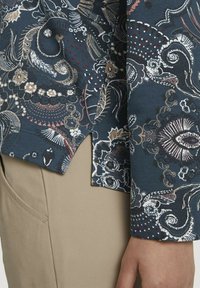 Camicia blu scuro con ornati floreali e paisley intricati in bianco, rosa e oro. Presenta un orlo curvo e una texture del tessuto morbida.