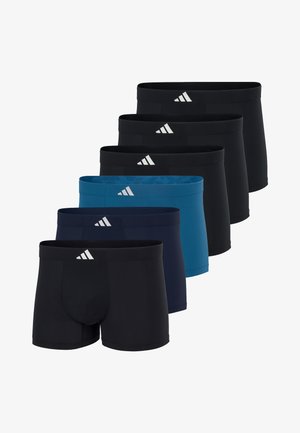 Athletischer Mann steht barfuß, trägt blaue, eng anliegende Boxer-Shorts mit einem kleinen weißen Logo am Bund, vor einem schlichten hellen Hintergrund.