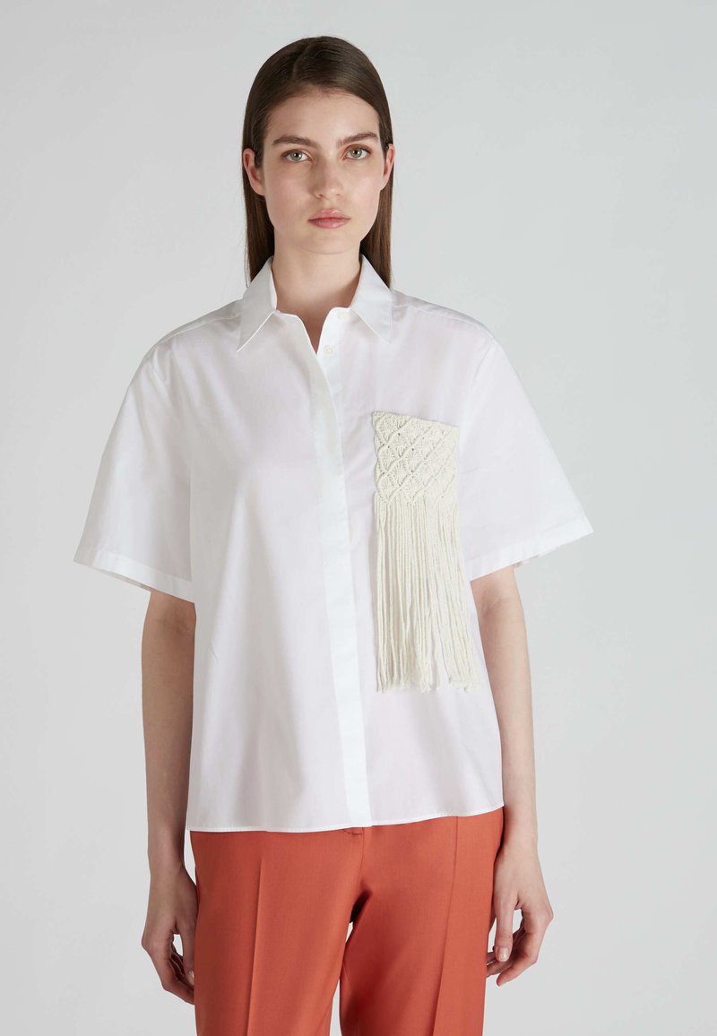 Camicetta bianca a maniche corte con colletto, caratterizzata da una tasca frangiata in avorio testurizzato sul lato sinistro; vestibilità sartoriale.