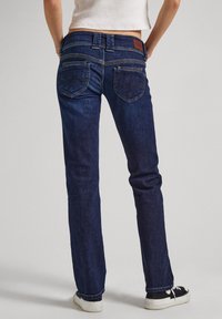 Pessoa vestindo jeans retos azul-escuro e ténis preto e branco, de pé com as mãos nos quadris contra um fundo neutro.