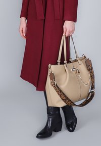 Borsa a tracolla in pelle beige con finitura testurizzata, dotata di doppi manici e tracolla decorata in marrone e nero, abbinata a un cappotto bordeaux.