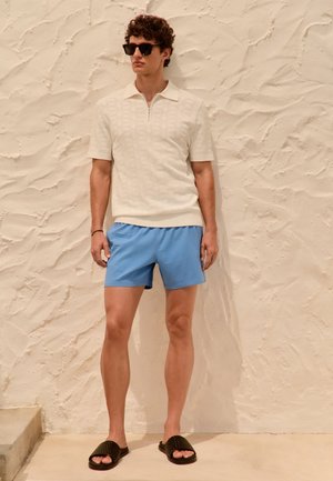 Ung mand iført tekstureret cremefarvet kortærmet polo, lyseblå shorts, sorte slidesandaler og solbriller står mod en tekstureret beige væg.