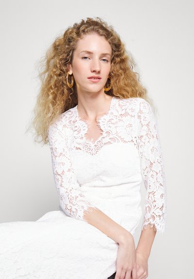 Kvinne med langt krøllete blondt hår iført gull hoops øreringer og en hvit blonderkjole med trekvartlange ermer, poserer mot en ensfarget bakgrunn.