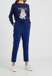 Felpa blu marina con grafica di un orso e testo "POLO", abbinata a pantaloni blu con strisce laterali e tasche. Sneakers bianche completano l'outfit.