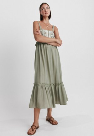 Junge Frau mit kurzen braunen Haaren, die ein ärmelloses olivgrünes Midikleid mit Stickerei auf dem Oberteil und braune Sandalen trägt.