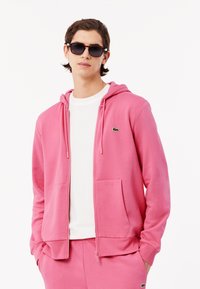 Haut à fermeture éclair rose avec cordon de serrage, poches avant et logo brodé. Porté sur une chemise blanche, associé à un pantalon de survêtement rose assorti.