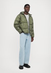 Bărbat îmbrăcat cu o jachetă puffer cu model în verde măsliniu peste o cămașă gri, blugi largi bleu deschis și pantofi negri, stând cu mâinile în buzunare.