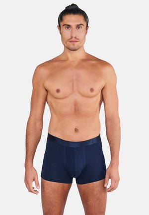 Skjorteløs mand med mørkt hår i en knold iført navy blå boxershorts, stående mod en ensfarvet lys baggrund.