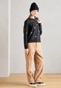 Felpa nera con ricami, dotata di una targhetta in pelle quadrata, abbinata a pantaloni cargo beige. Il modello indossa un berretto di maglia nero e sneakers.