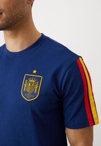 Μπλε βαμβακερό t-shirt με κίτρινο έμβλημα στο στήθος και κόκκινες και κίτρινες ρίγες στα μανίκια. Σχεδίαση με κοντά μανίκια.