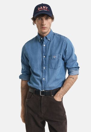 Camisa de mezclilla azul con botones, bolsillo en el pecho, mangas enrolladas y pantalones marrones oscuros, complementada con una gorra navy con texto rojo.