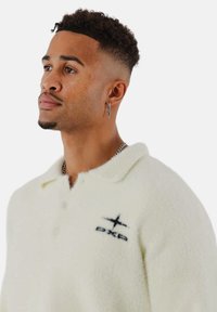 Flauschiges cremefarbenes Polo-Shirt mit Kragen, drei Knöpfen und einem schwarzen Logo auf der linken Brust. Strukturiertes Gewebe und entspannte Passform.